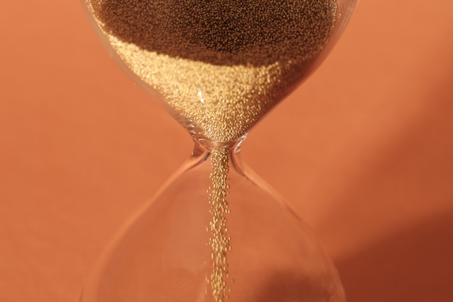 Close-up of golden sand flowing through the narrow center of an hourglass against orange background.